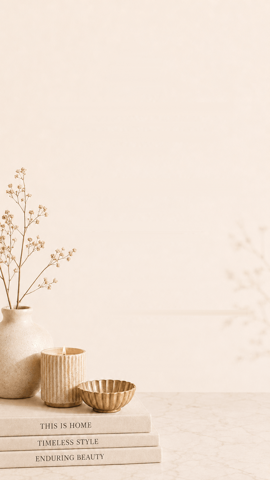 A ceramic vase of dried gypsophila, a ribbed candle and a brass bowl beside a stack of design books on a marble surface.